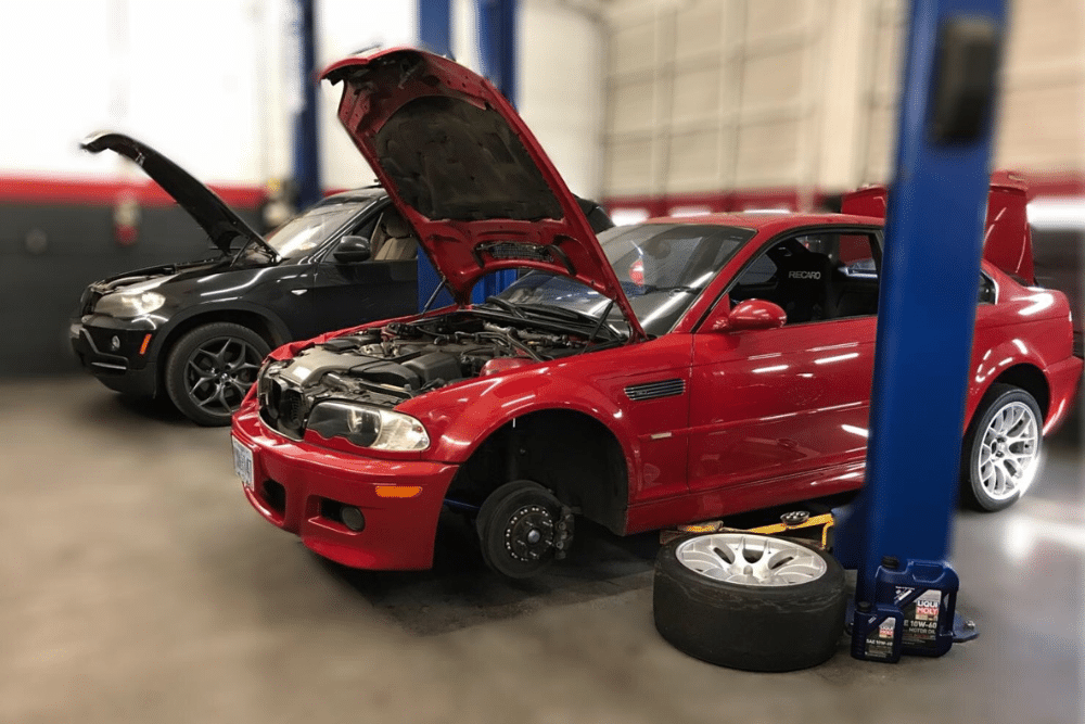 Car Maintenance, auto repair in Bend, OR at Mountain View Imports. Two cars in an auto repair shop with hoods open, a black SUV and a red sports coupe on a lift. The red car has one front wheel removed, with the wheel and motor oil containers placed on the ground nearby.